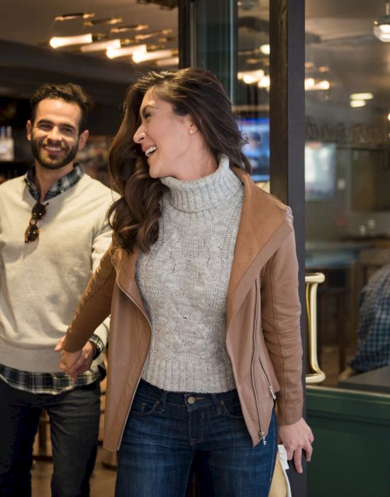 A smiling woman and man walk out of a cozy cafe. The ambiance inside appears warm, with people seated and lights glowing softly.