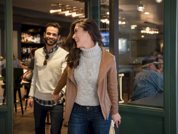 A smiling woman and man walk out of a cozy cafe. The ambiance inside appears warm, with people seated and lights glowing softly.