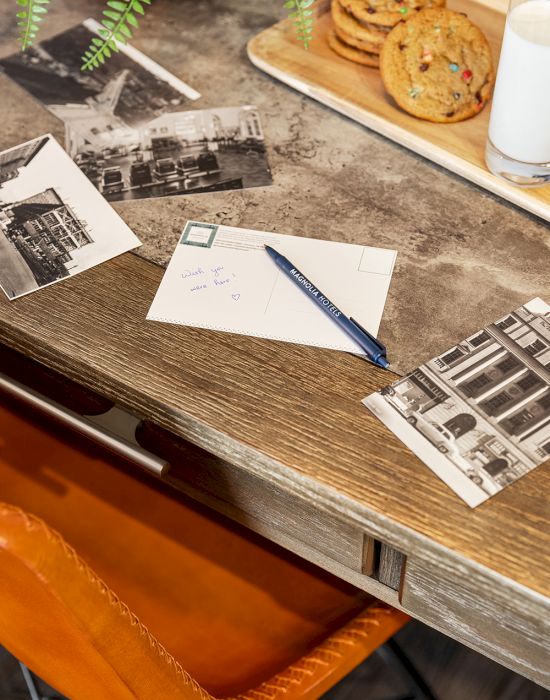 A desk with postcards, a pen, cookies, and milk on a wooden surface, next to an orange chair.