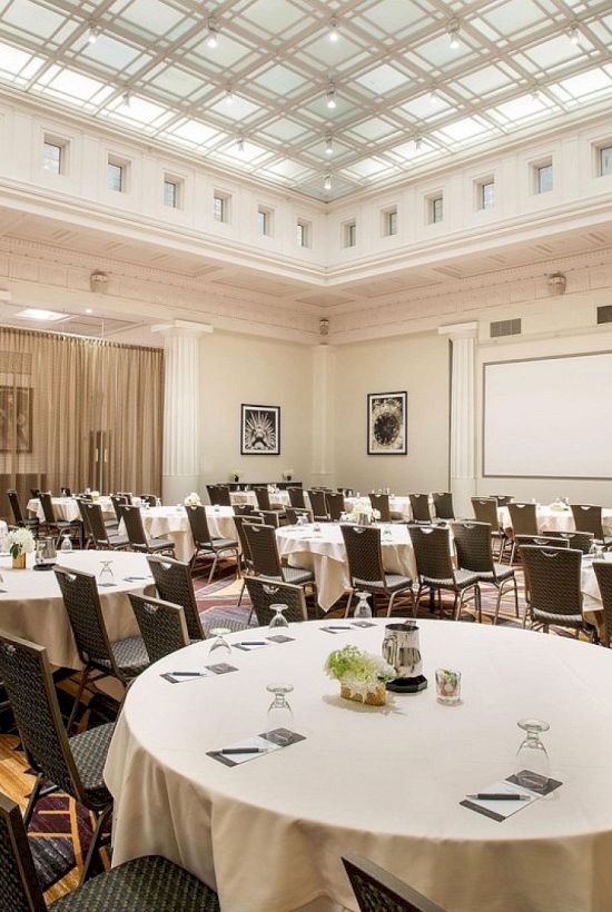 The image shows an elegant conference room with round tables and chairs arranged, a projector screen, and decorative ceiling.