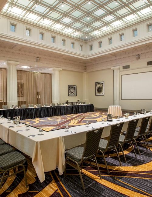 A conference room set up in a U-shape with chairs, tables, a projector screen, and a patterned carpet under a large, illuminated ceiling.