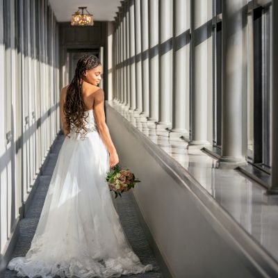 A person in a bridal gown stands in a sunlit corridor, holding a bouquet, with large windows along one side and modern lights above.