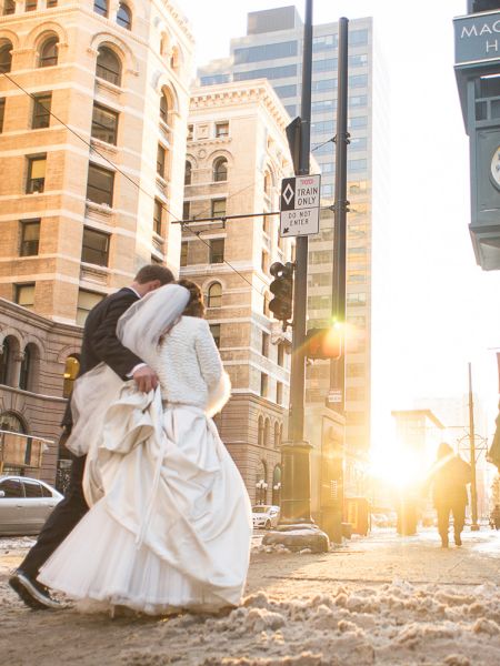 A couple in wedding attire embraces on a city street. The sun sets behind them near a building with a 