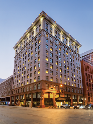 A multi-story historic building with lights on, corner view, surrounded by cars, at dusk.