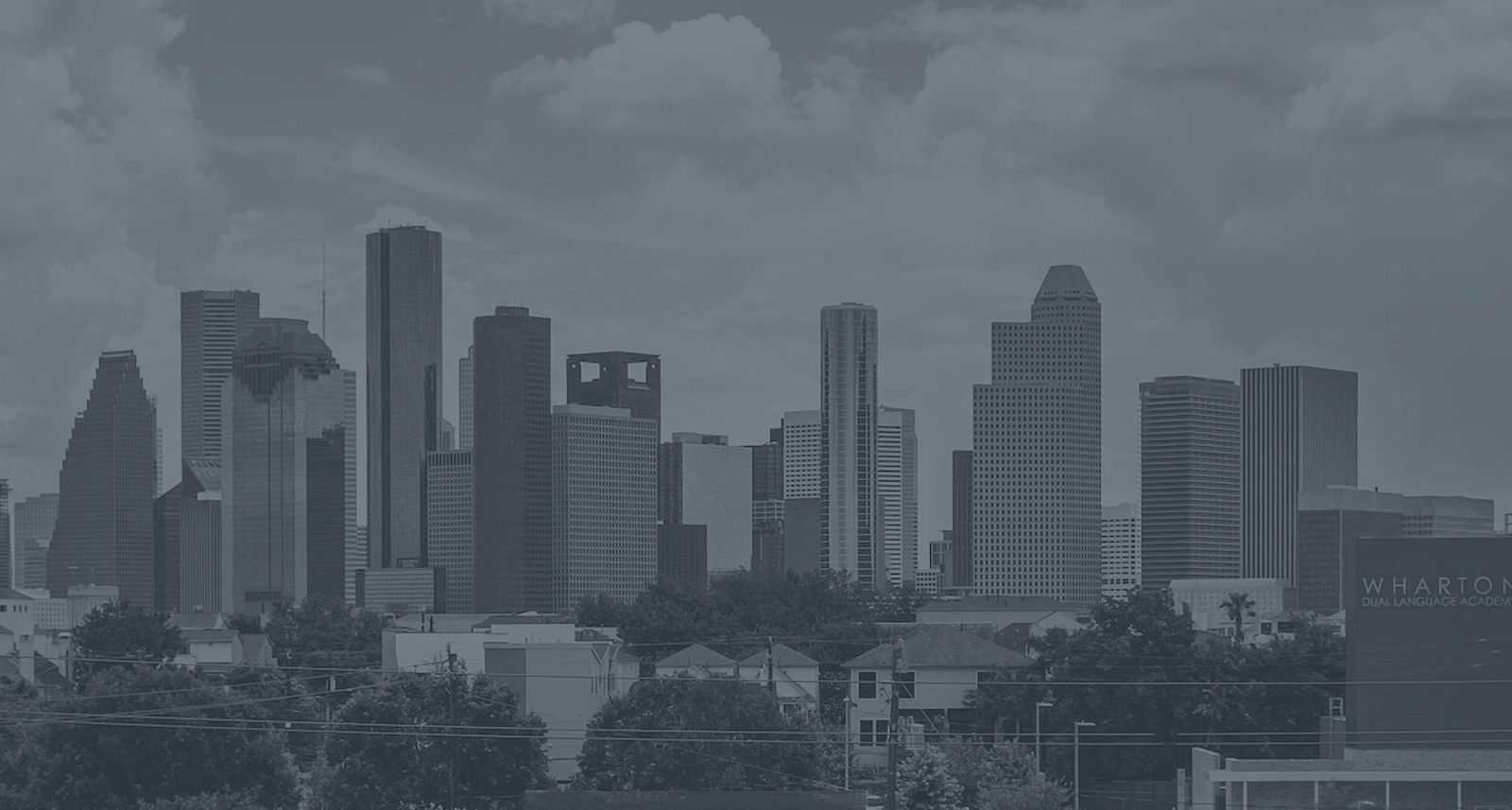 A city skyline featuring various skyscrapers under a partly cloudy sky, with trees and a building in the foreground.