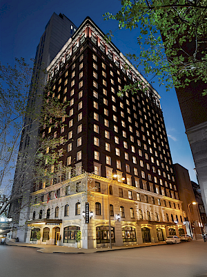 The image shows a tall, elegantly-lit building on a street corner at dusk, featuring many windows and a classic architectural style.