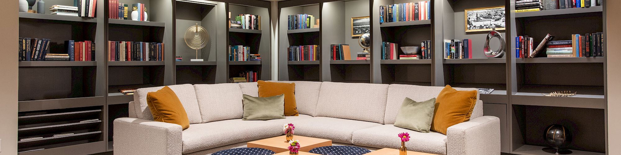 A cozy library lounge with a sectional sofa, bookshelves, a patterned carpet, and two small tables with decorative vases.