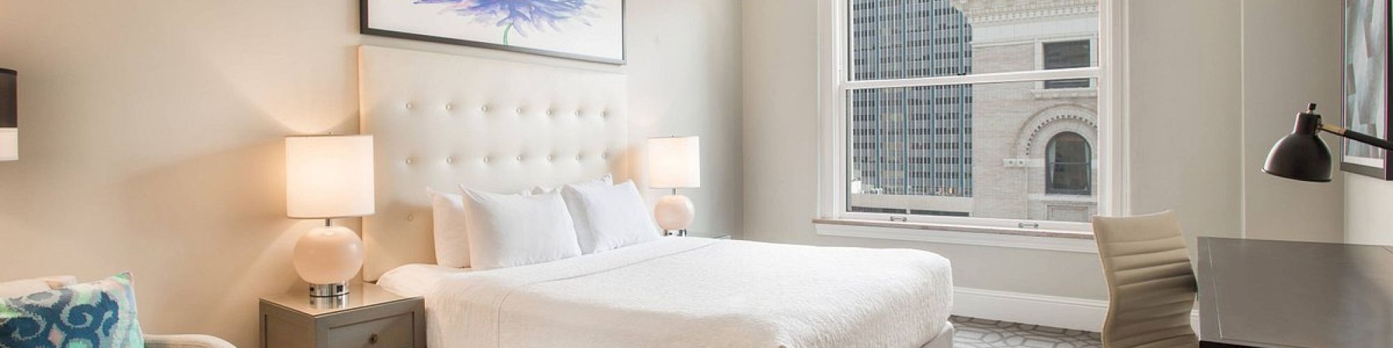 The image shows a modern hotel room with a bed, armchair, desk, and window view. The decor is simple and elegant.