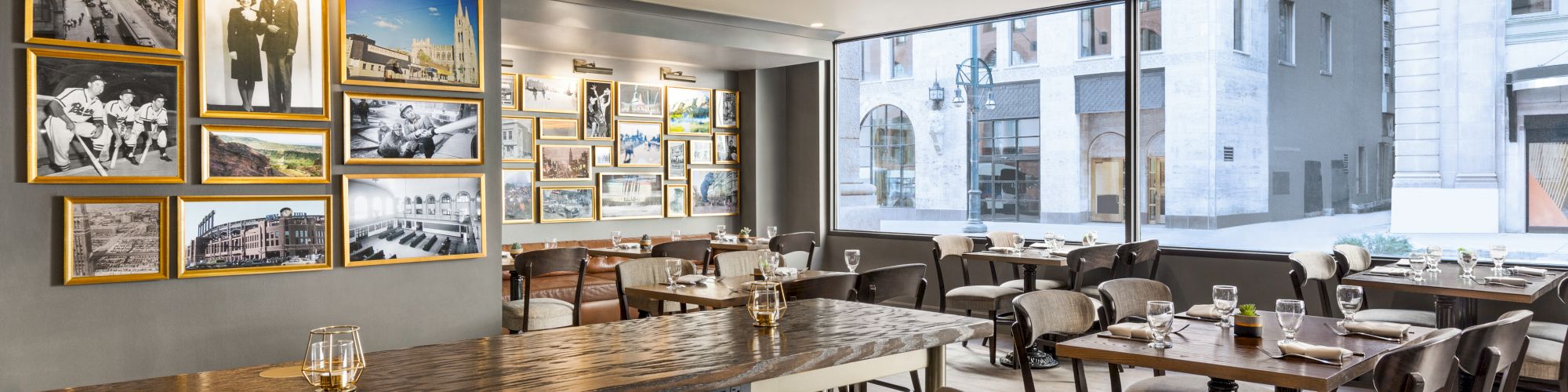 A modern restaurant with framed photos on the wall, wooden tables, and cushioned chairs, featuring large windows with street view.