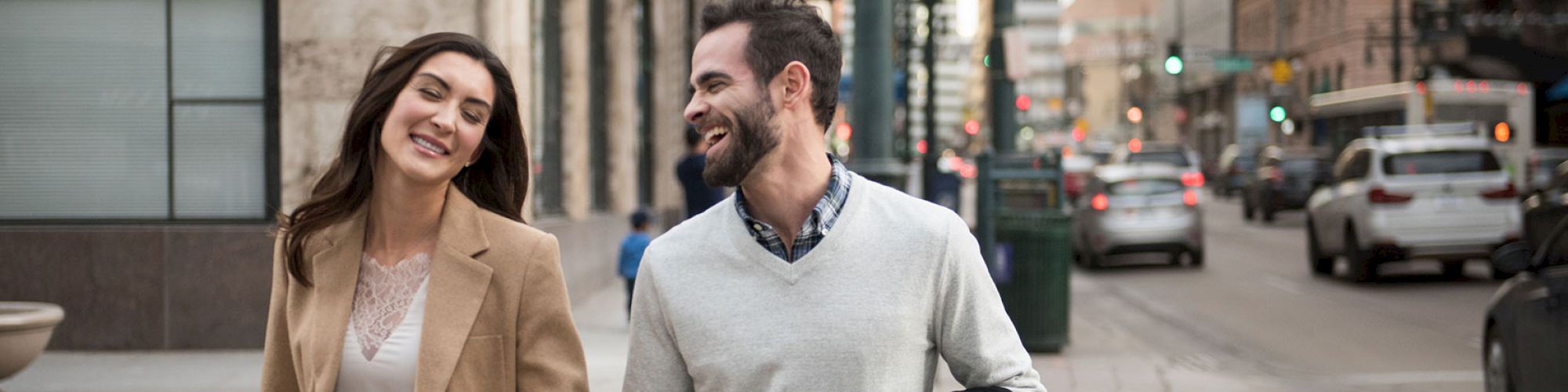 A couple is holding hands and smiling while walking on a city street lined with buildings and cars.