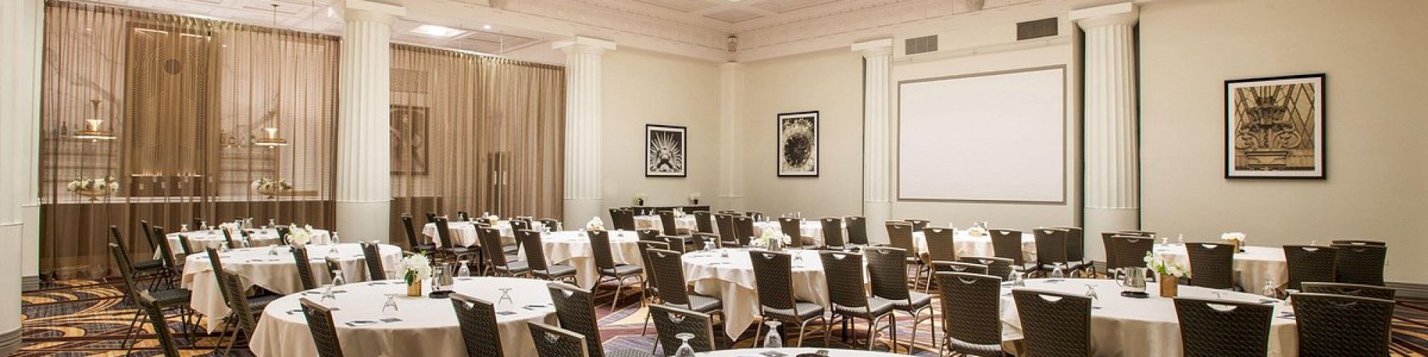 The image shows a conference room set up with round tables, chairs, a large screen, and elegant lighting from a skylight ceiling.