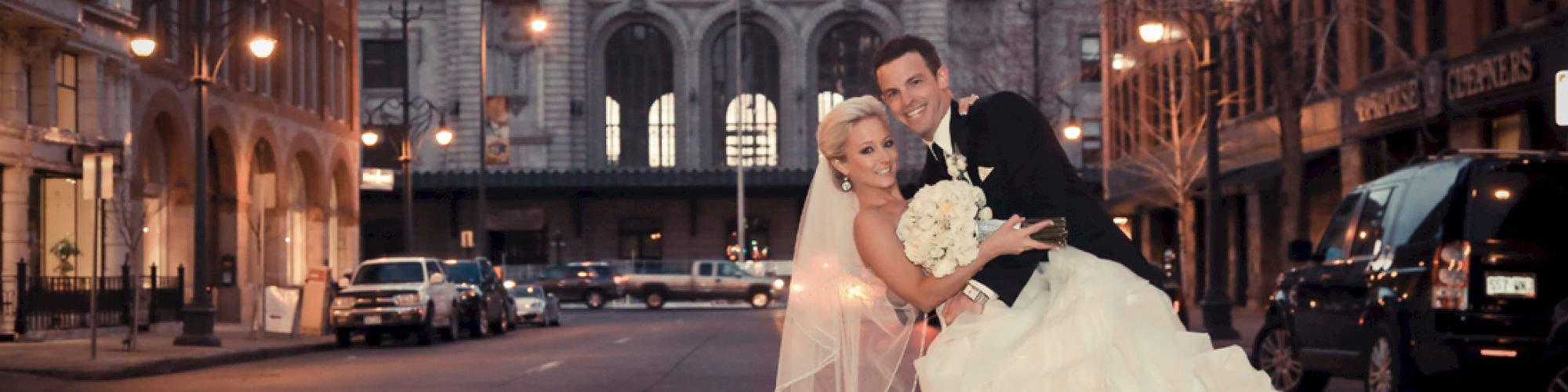 A couple in wedding attire poses joyfully in front of Union Station on a city street with an elegant, urban backdrop.