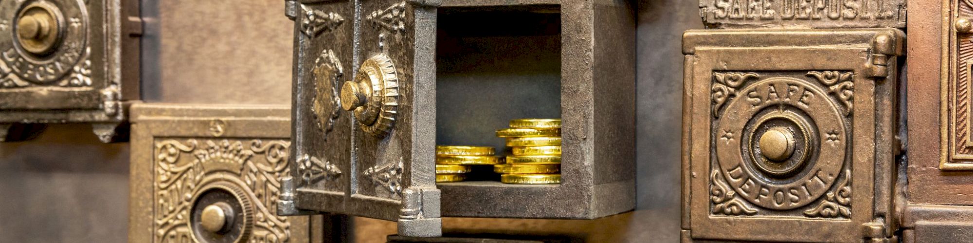 The image shows a wall with vintage safe deposit boxes. One box is open, revealing stacks of gold coins inside.