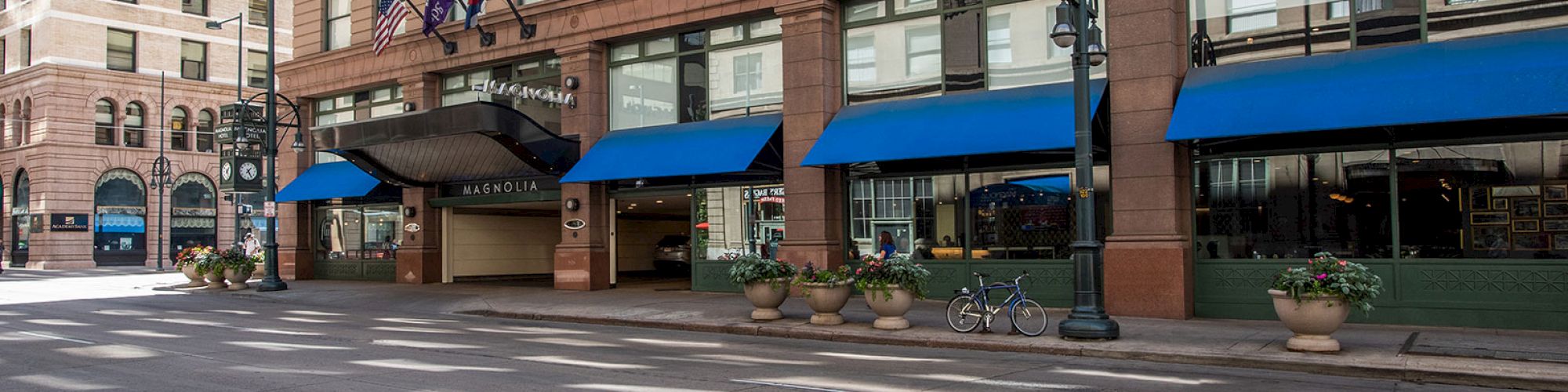 The image shows a street view of a large building with blue awnings, flags, and potted plants outside. It appears to be a hotel entrance.