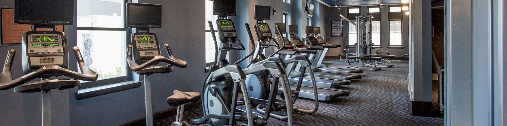 A gym with stationary bikes and treadmills lined up, equipped with screens, under a ceiling with spotlights and large windows.