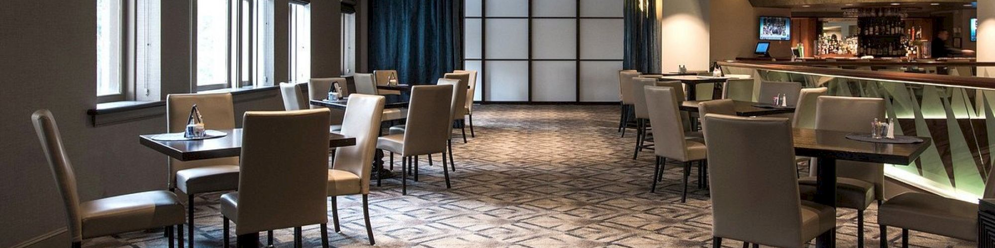 The image shows an empty, modern restaurant with tables and chairs arranged neatly, featuring carpeted flooring and a bar area in the background.