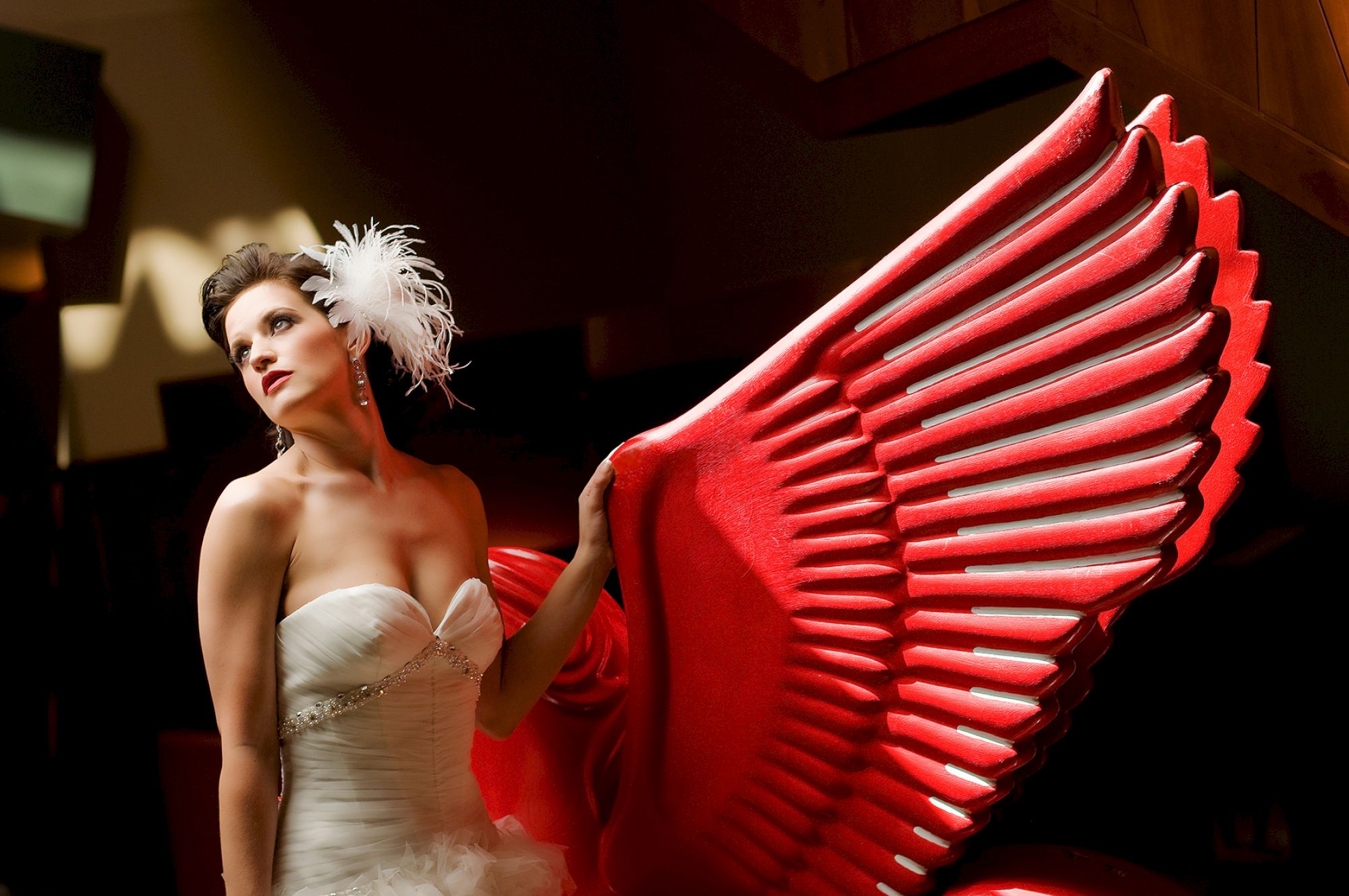 A woman in a white dress, adorned with feathers, poses beside large red wings in a dramatic setting.