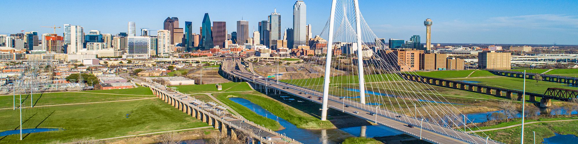 The image shows a city skyline with modern skyscrapers and a distinctive cable-stayed bridge, set against a clear blue sky.