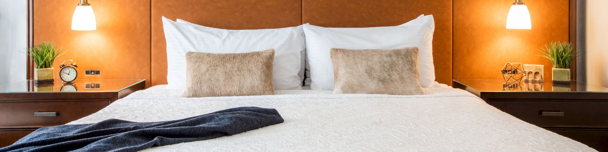 A neatly made bed with white bedding, brown headboard, bedside lamps, decorative pillows, a blue throw, and framed art on the headboard.