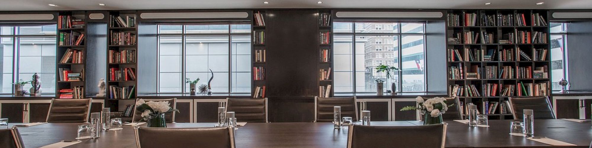 A modern conference room with a large table, surrounded by chairs, bookshelves, and decorative plants under stylish lighting fixtures.