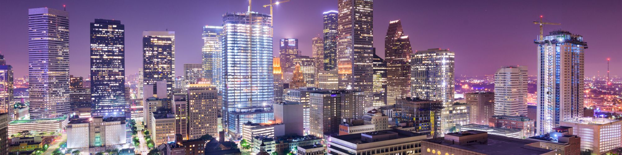 A vibrant city skyline at night with illuminated skyscrapers, construction cranes, and city lights under a colorful sky.