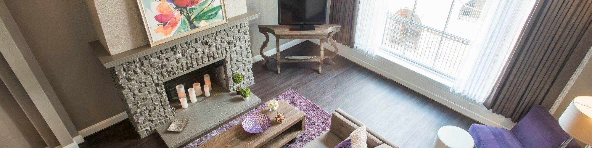 A modern living room with a stone fireplace, colorful painting, purple accents, sofa, TV, large window with curtains, and wooden flooring.