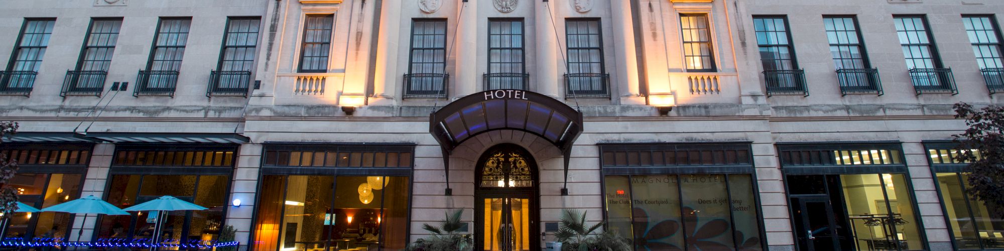 The image shows a four-story building with flags, lit in the evening. It appears to be a hotel with an entrance at the center.