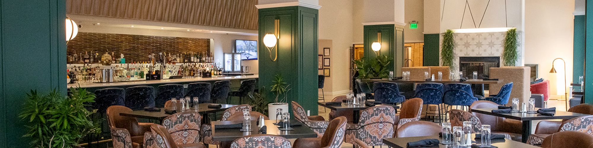 The image shows a stylish restaurant interior with patterned chairs, wooden tables, and a well-lit bar, surrounded by modern decor and plants.