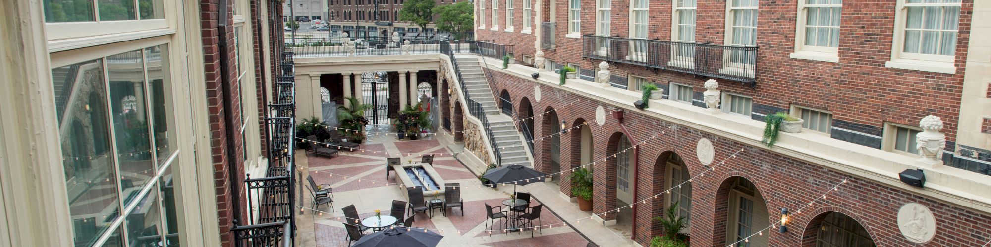 A courtyard with tables, chairs, and umbrellas, surrounded by a multi-story brick building with large windows and arches in view.