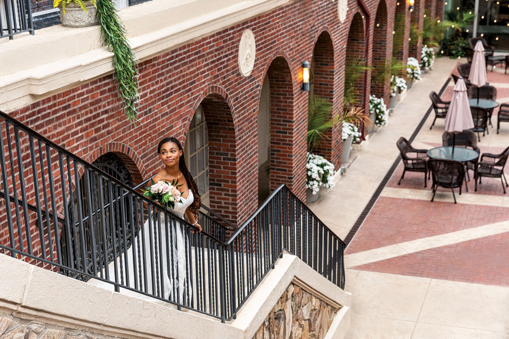 A person in a white outfit holding a bouquet stands on stairs near a brick building, overlooking a courtyard with tables and chairs.