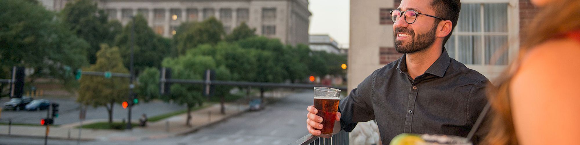 Two people are enjoying drinks on a balcony with a cityscape in the background, featuring a prominent building and street view.