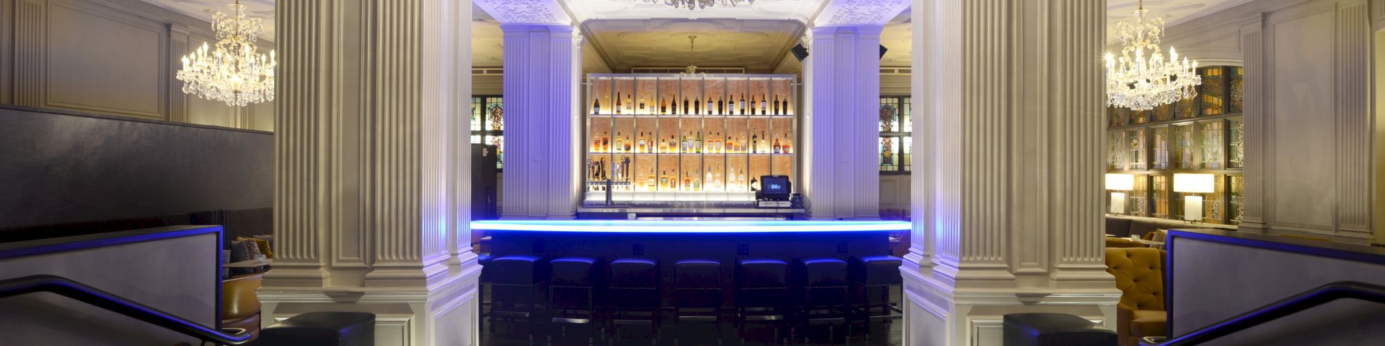 The image shows an elegant bar with chandeliers, mood lighting, and a blue-lit counter surrounded by stools and decorative columns.