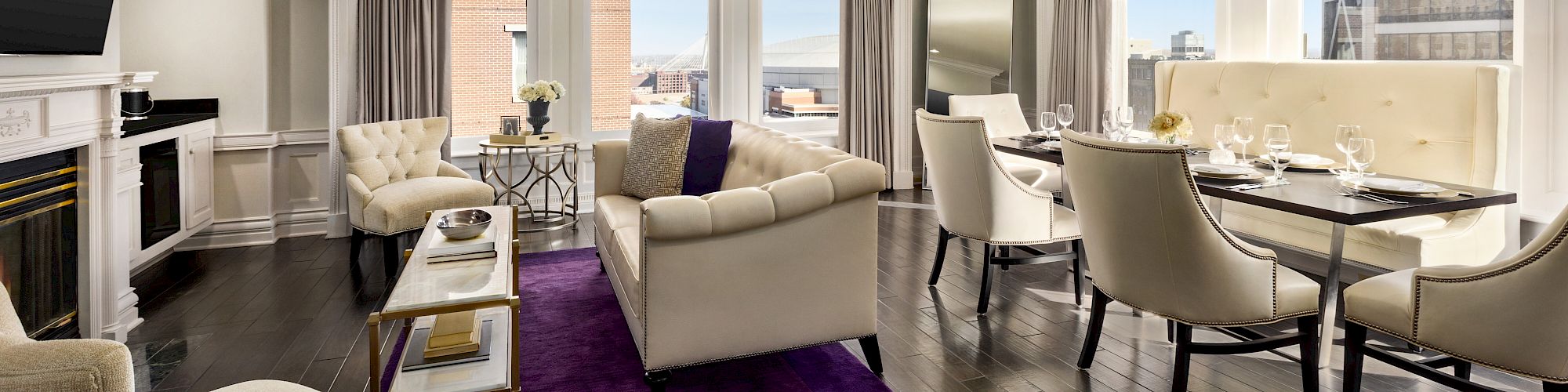 A modern, stylish living room with cream sofas, a purple rug, dining table, and large windows revealing a scenic view outside.