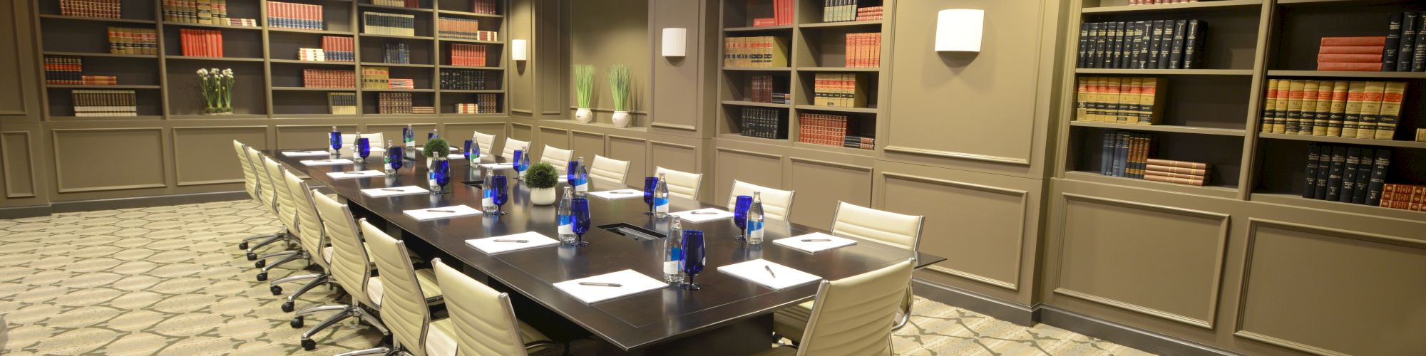 A conference room with a long table, chairs, and bookshelves filled with books. There are water bottles and papers on the table.