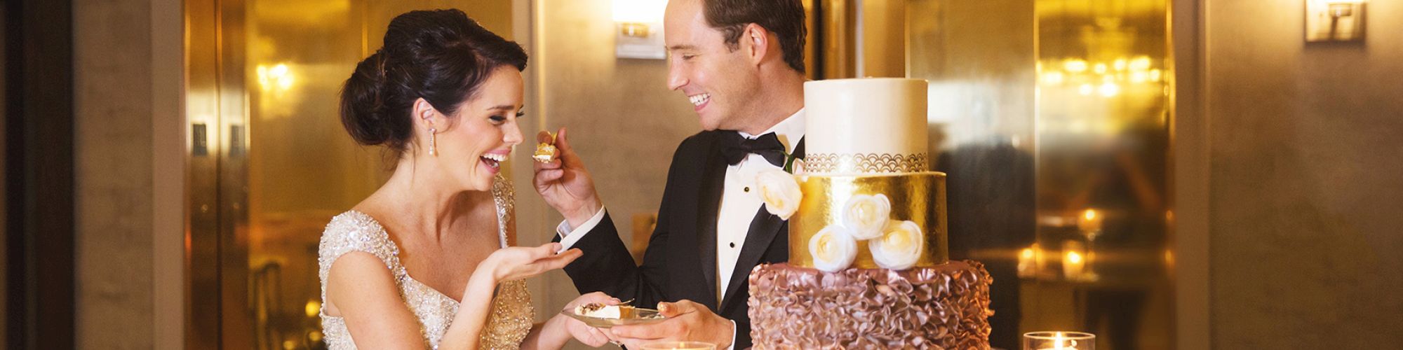 A couple, dressed in wedding attire, is enjoying cake together in a warmly lit setting with candles on the table.