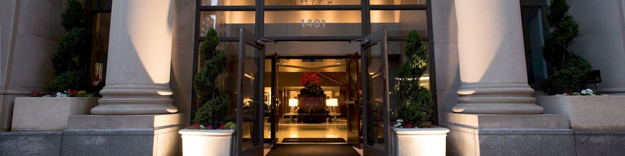 The image shows the entrance of a modern building with large glass doors, flanked by tall columns and planters, and lit with warm lighting.