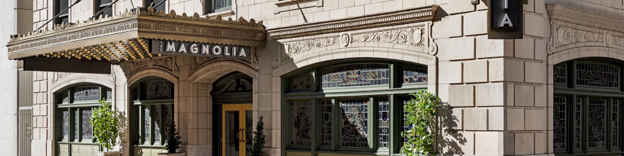 The image shows the exterior of The Magnolia Hotel with flags on the facade and a decorative canopy above the entrance.