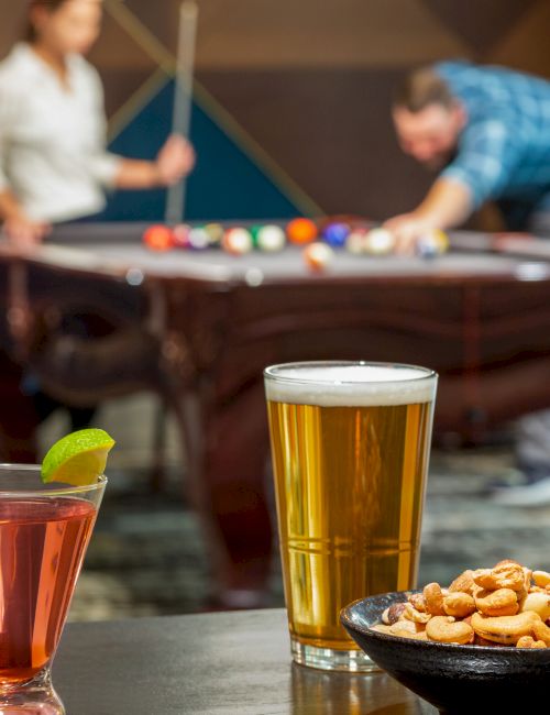 Drinks and snacks are on a table in the foreground, with two people playing pool in the blurry background.