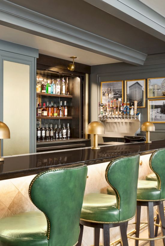 A stylish bar interior with green leather stools, a sleek countertop, bottles on display, and framed pictures on the wall.