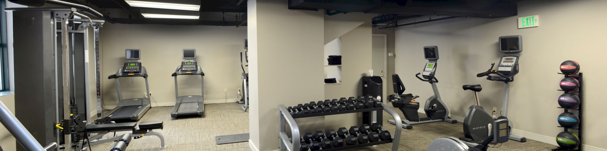 The image shows a gym with treadmills, stationary bikes, weights, and exercise benches in a carpeted room with bright lighting.