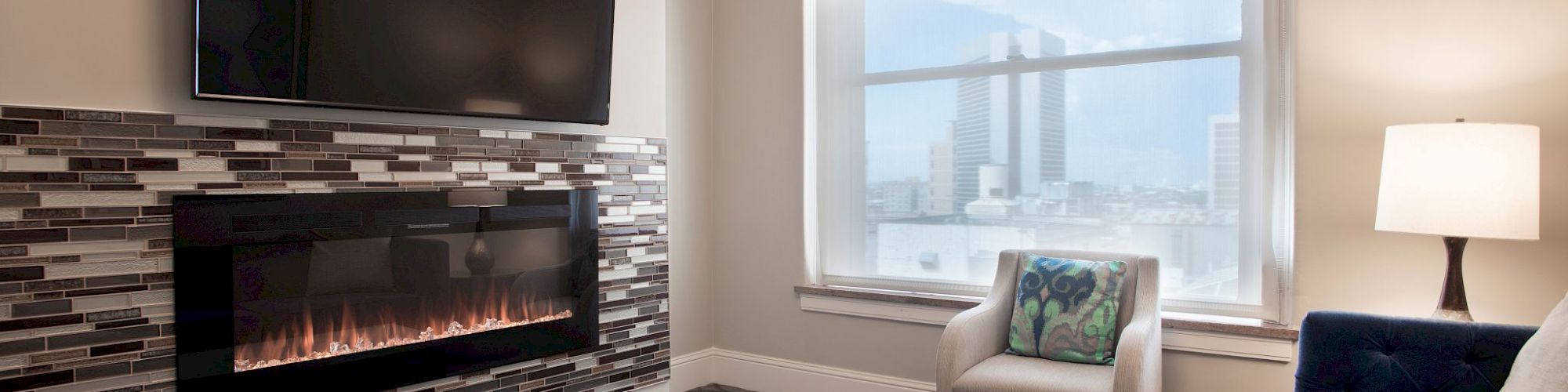 A modern living room with a wall-mounted TV, electric fireplace, sofa, armchair, coffee table, and window with a city view.