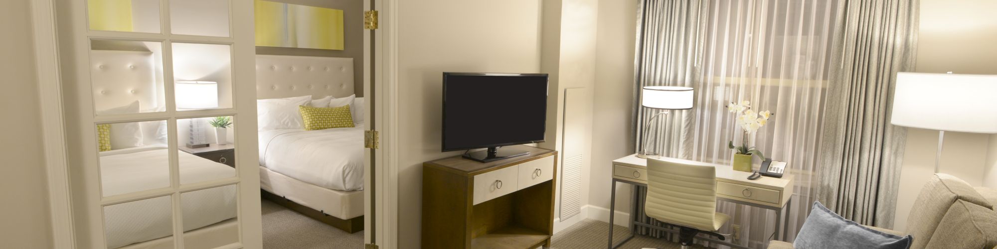 The image shows a hotel suite with a living area, TV, desk, and a bedroom with a bed, separated by French doors, in neutral tones.