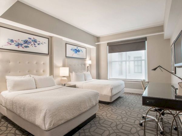 A modern hotel room with two double beds, decorative wall art, a desk, and a lamp, featuring a large window with a rolled-up blind.