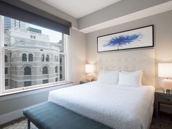 A modern hotel room with a large bed, two lamps, a colorful wall art piece, and a view of historic buildings through the window.