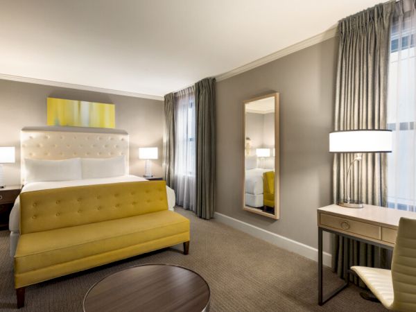 A modern hotel room with a bed, yellow bench, desk, chair, mirror, and lamps; neutral tones and a window with curtains complete the decor.