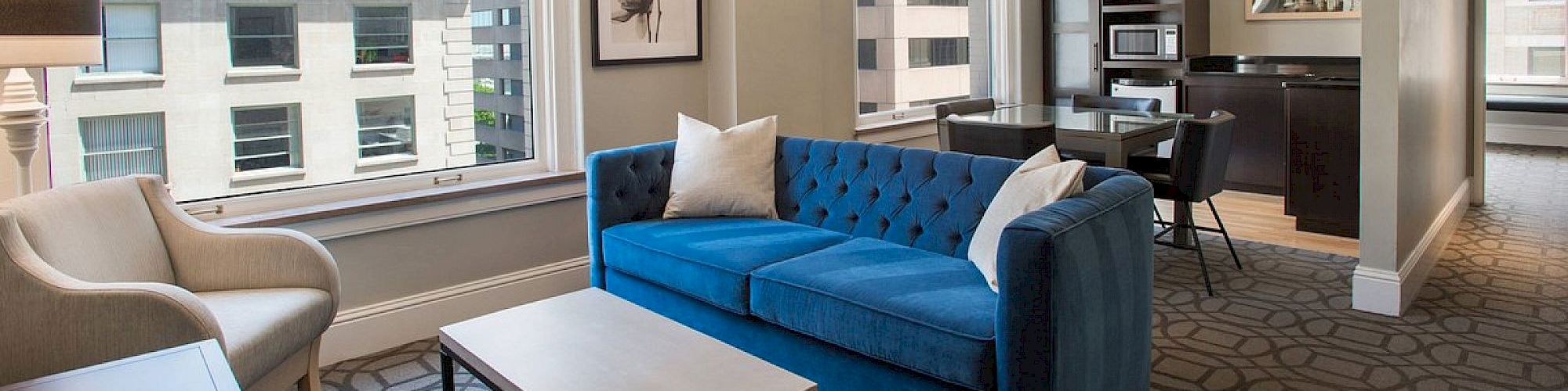 A modern living room with a blue sofa, armchair, coffee table, and dining area. Large windows offer city views, enhancing the space.