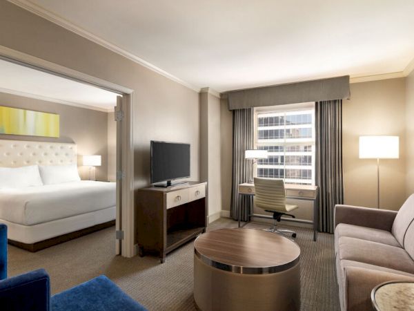 The image shows a hotel room with a bed, TV, desk, sofa, and a window with city view, decorated in neutral tones.