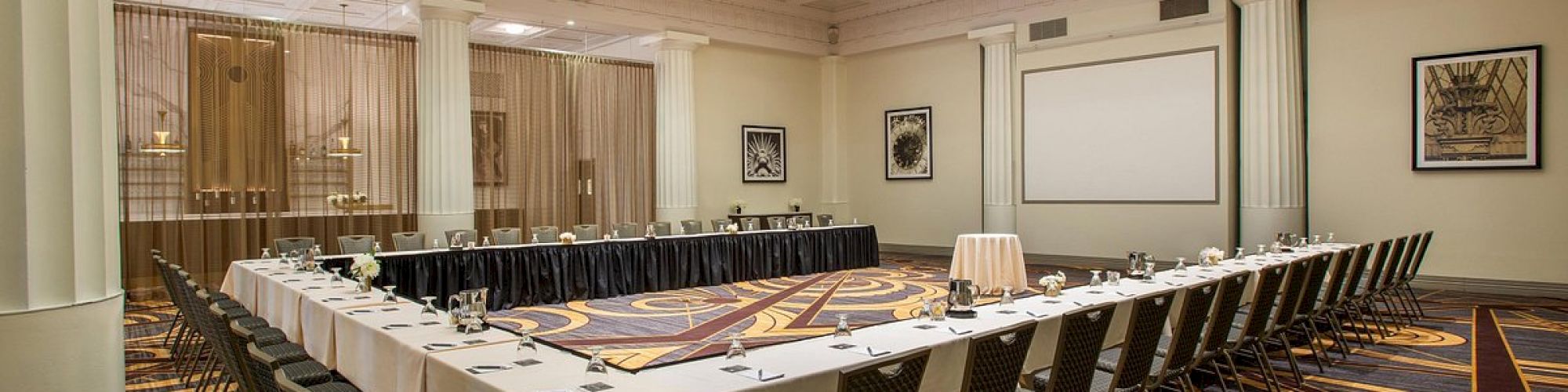 A large conference room with a U-shaped table setup, chairs, and a projector screen. The space has a patterned carpet and ceiling lights.