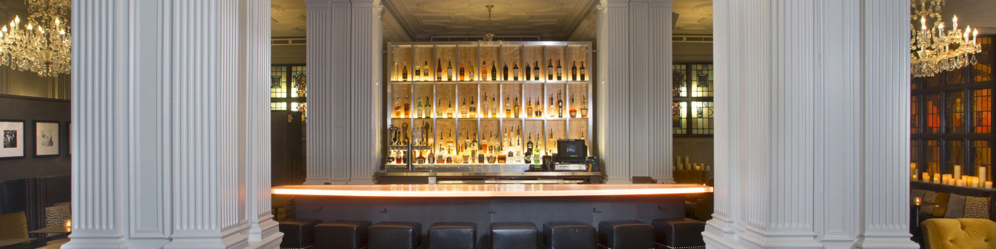 The image shows an elegant bar with a chandelier, high ceiling, and columns, featuring bottles displayed behind the counter and several seats.