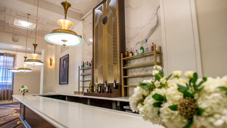 The image shows an elegant bar interior with decorative lighting, flower arrangements, and a stylish shelving unit with bottles.